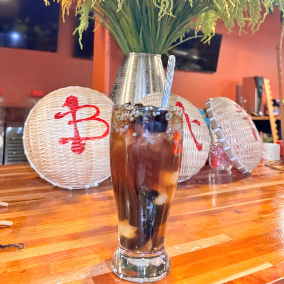 A Vietnamese dessert drink with dark jelly, white fruit, and seeds in a tall glass on a wooden counter.