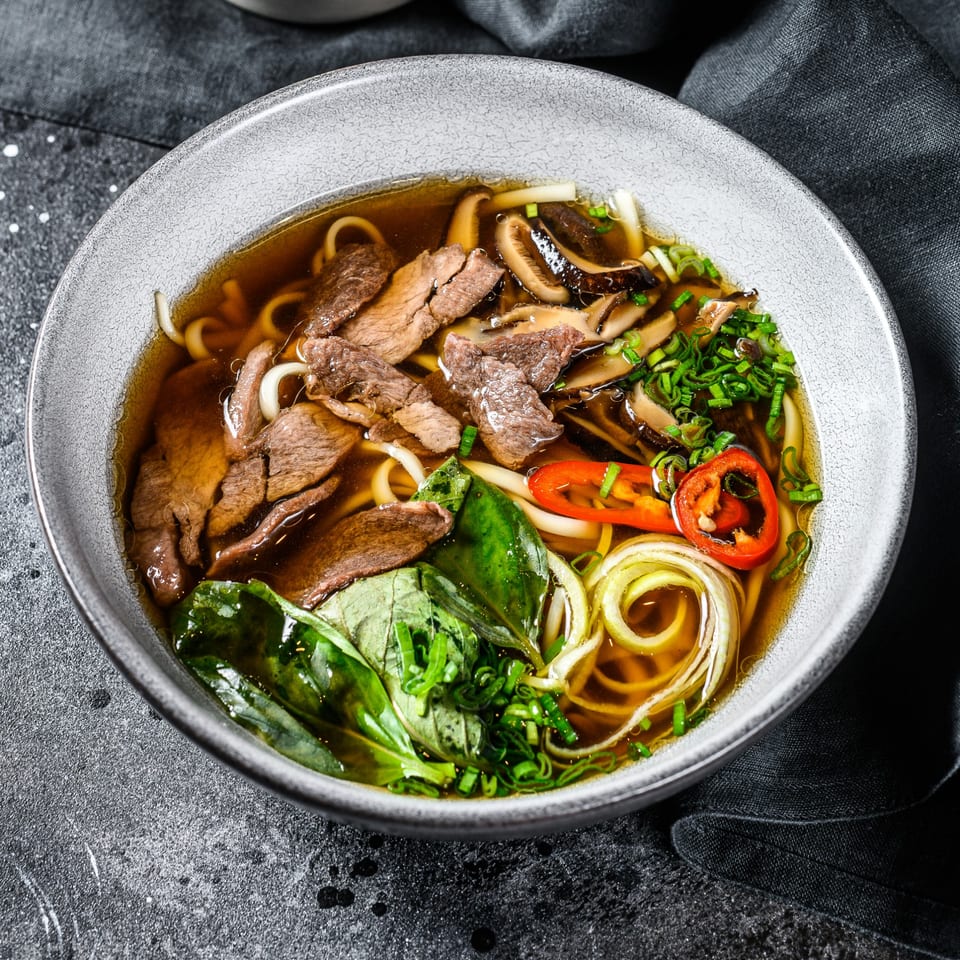 Vietnamese pho with beef, mushrooms, herbs, and chili in a gray bowl on a dark background.